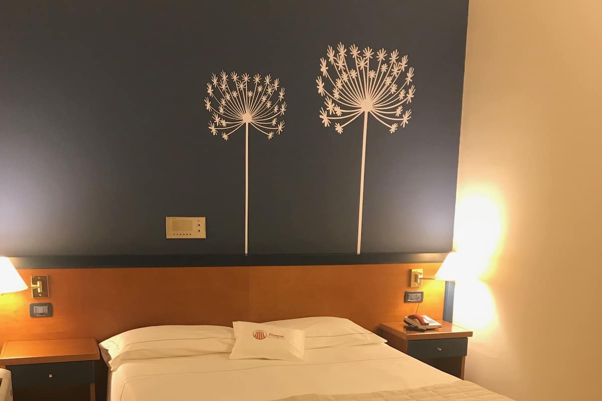 A hotel room with a bed, two bedside tables, a nightstand, a wall with two decorative tree designs, and a wall-mounted thermostat.