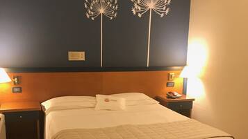 A hotel room with a bed, two bedside tables, a nightstand, a wall with two decorative tree designs, and a wall-mounted thermostat.