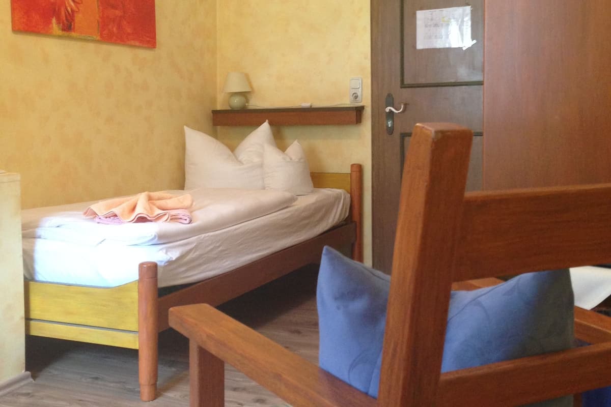Una habitación con una cama, una silla de madera con cojines, un armario de madera y una estantería con lámpara.