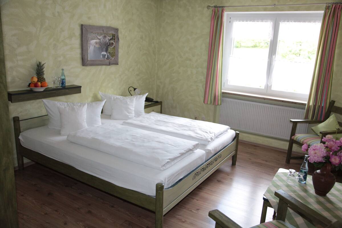 Una cama bien tendida con sábanas blancas, cabecera de madera, mesita de noche con una botella y un vaso, y una ventana con cortinas.