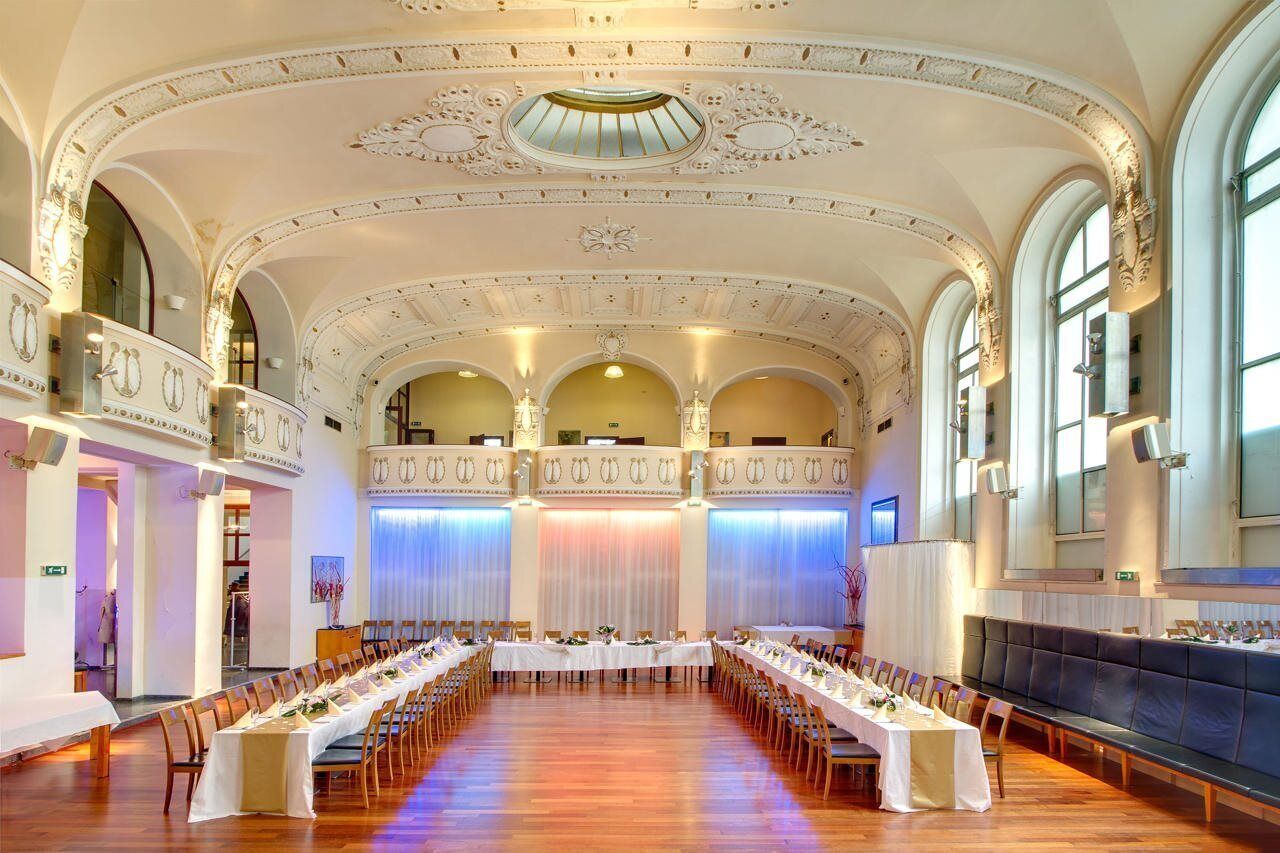 Un amplio salón de banquetes con largas mesas dispuestas para comer, techos altos con decoraciones ornamentales y grandes ventanales que permiten la entrada de luz natural.