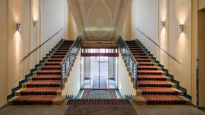 Una imponente escalera con descansillo central, flanqueada por paredes con luces y un techo decorativo.