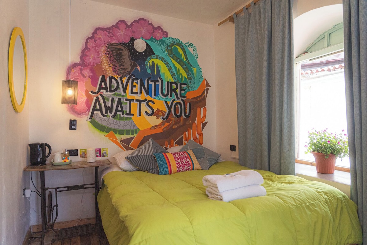 A room with a bed, a table with a kettle, a wall with a colorful mural, and a window with curtains.