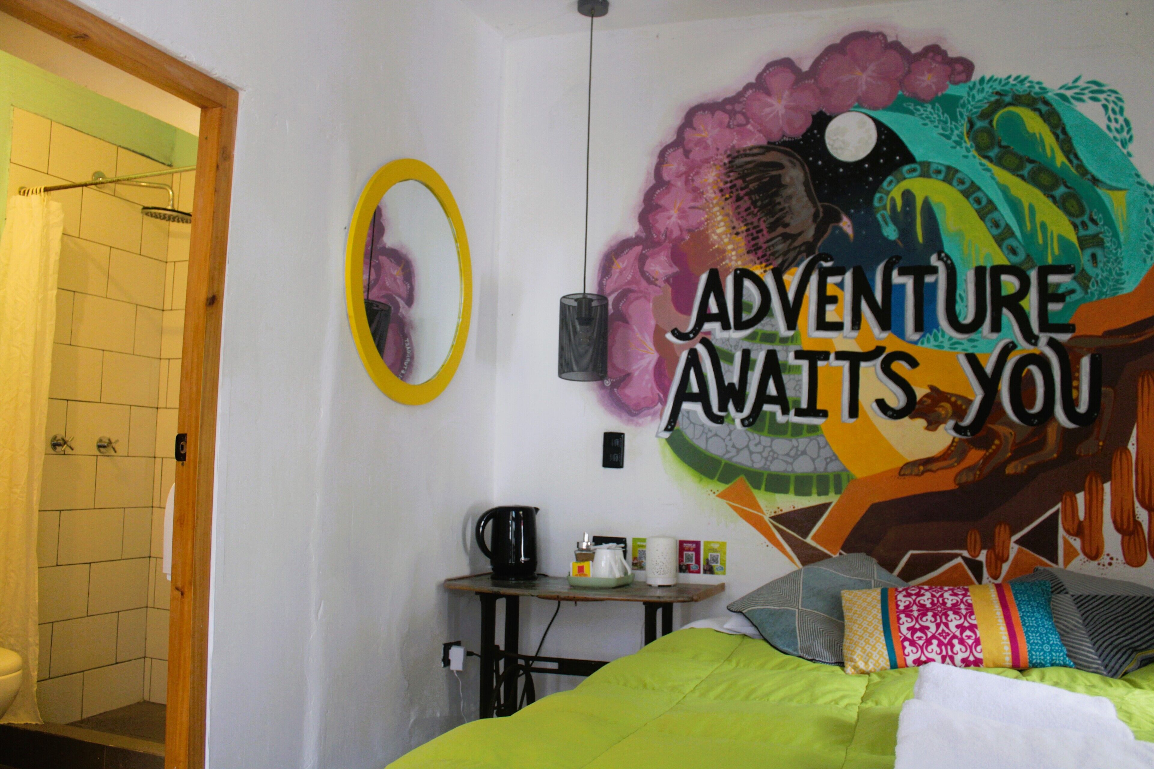 A room with a bed, a small table with a kettle, a mirror, and a wall with a colorful mural saying "ADVENTURE AWAITS YOU".