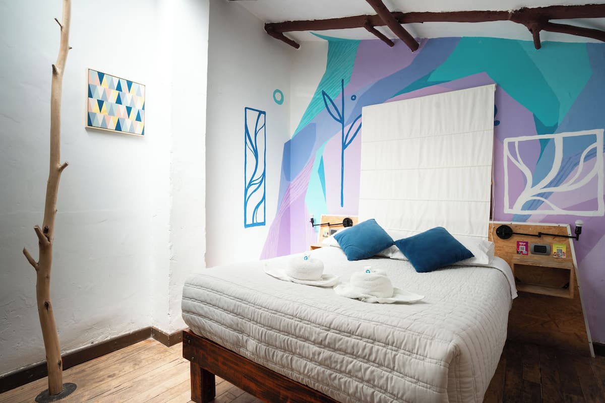 A bedroom with a bed, pillows, a bedside table, a wall with abstract art, and a wooden floor.