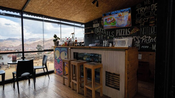 Un espace bar doté d’un plafond en bois, de grandes fenêtres offrant une vue sur les montagnes et d’un tableau noir affichant un menu proposant diverses boissons et plats.