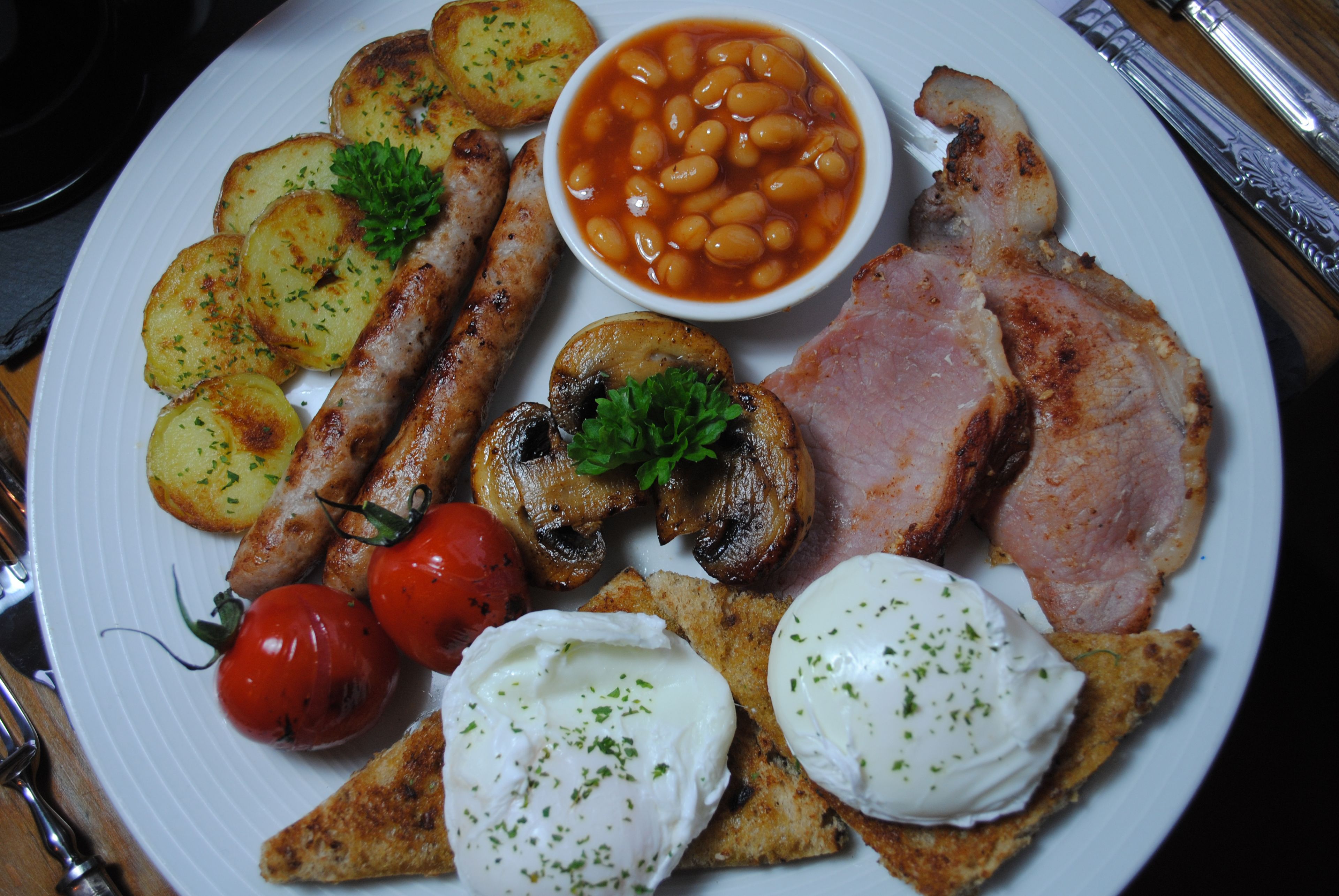 A plate of food including sausages, mushrooms, tomatoes, beans, ham, and poached eggs.