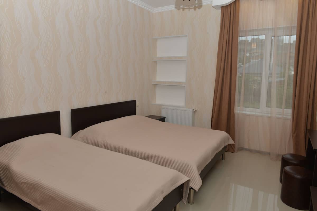A room with two beds, a radiator, a shelf unit, and a window with curtains.