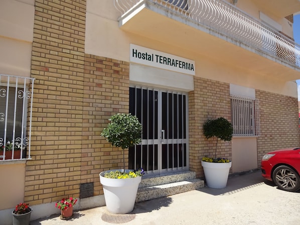 Un edificio con un letrero que dice "Hostal TERRAFERMA" y un coche rojo estacionado en la parte delantera. Hay plantas en macetas frente a la entrada.