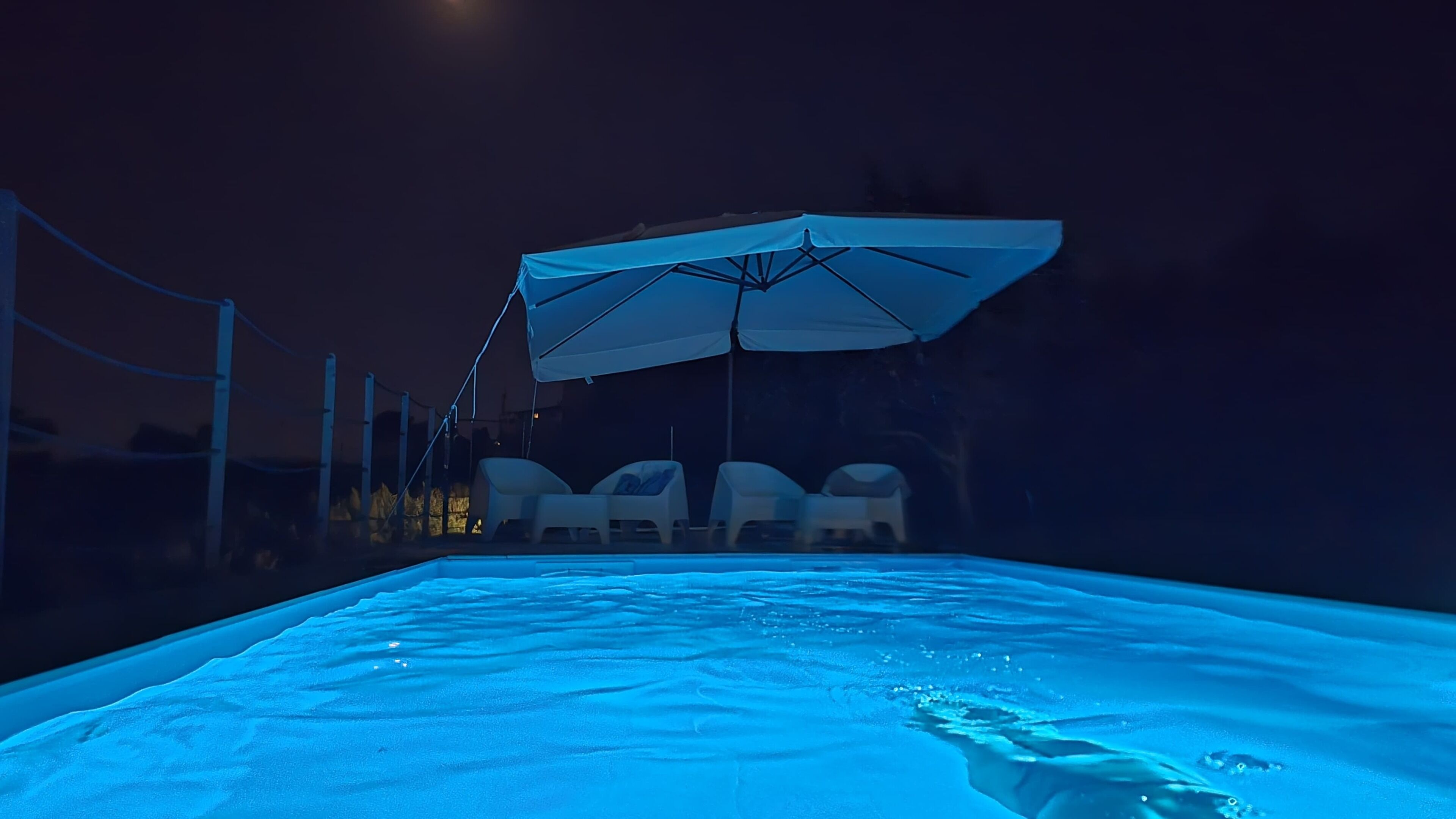 A swimming pool at night with a blue light. There are lounge chairs and umbrellas.