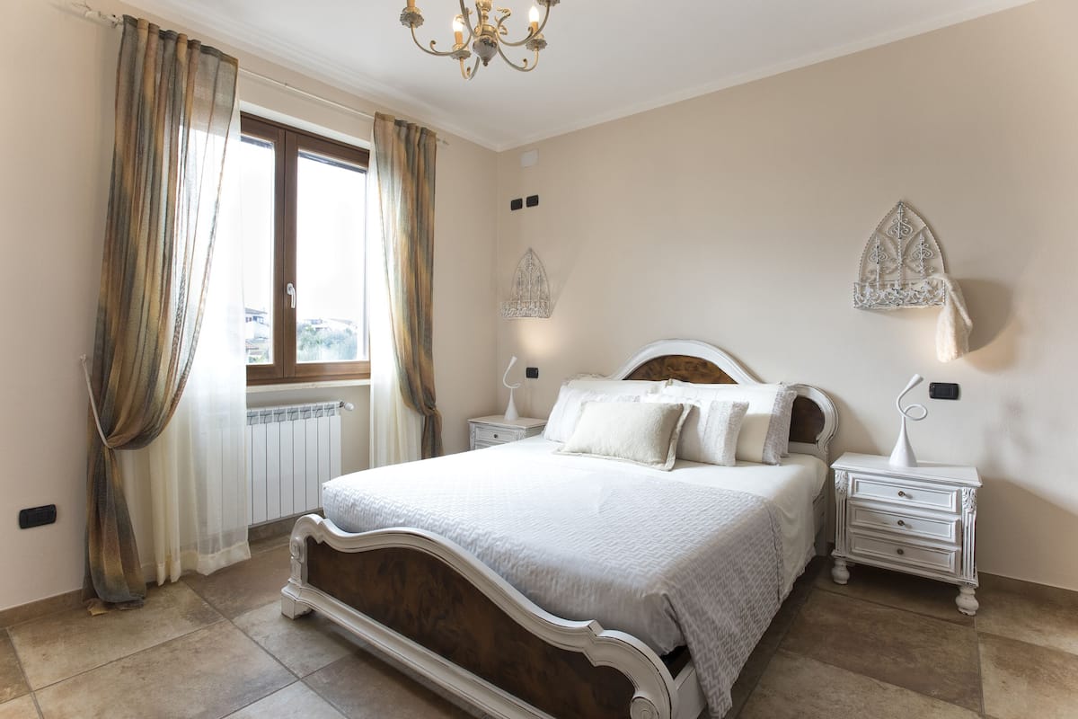 Une chambre à coucher avec un grand lit, deux tables de chevet, une fenêtre avec des rideaux et un lustre.
