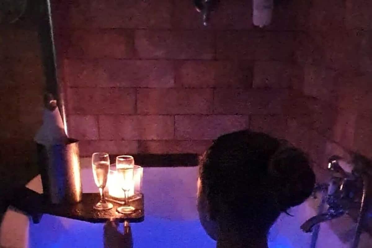 Une personne dans une baignoire, éclairée d’une lumière bleue, tenant un plateau avec des verres.