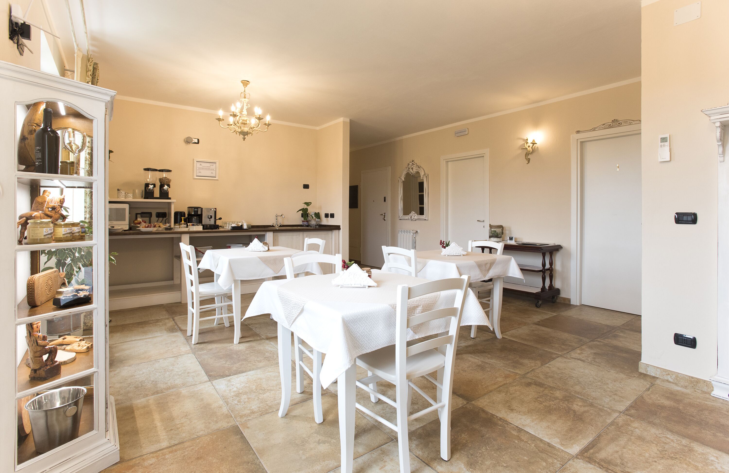 A dining area with white tables and chairs, a kitchen in the background, and a chandelier.
