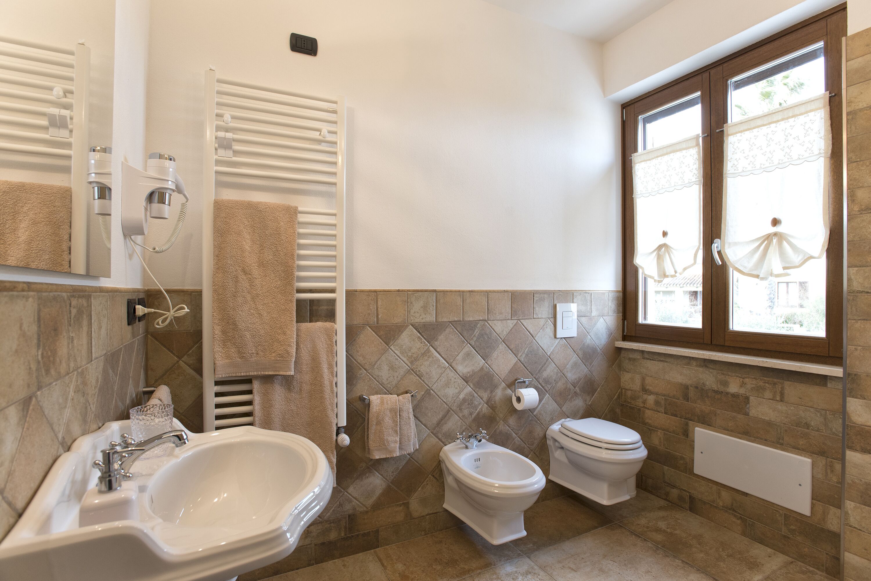 A bathroom with a sink, bidet, toilet, and a window with curtains.
