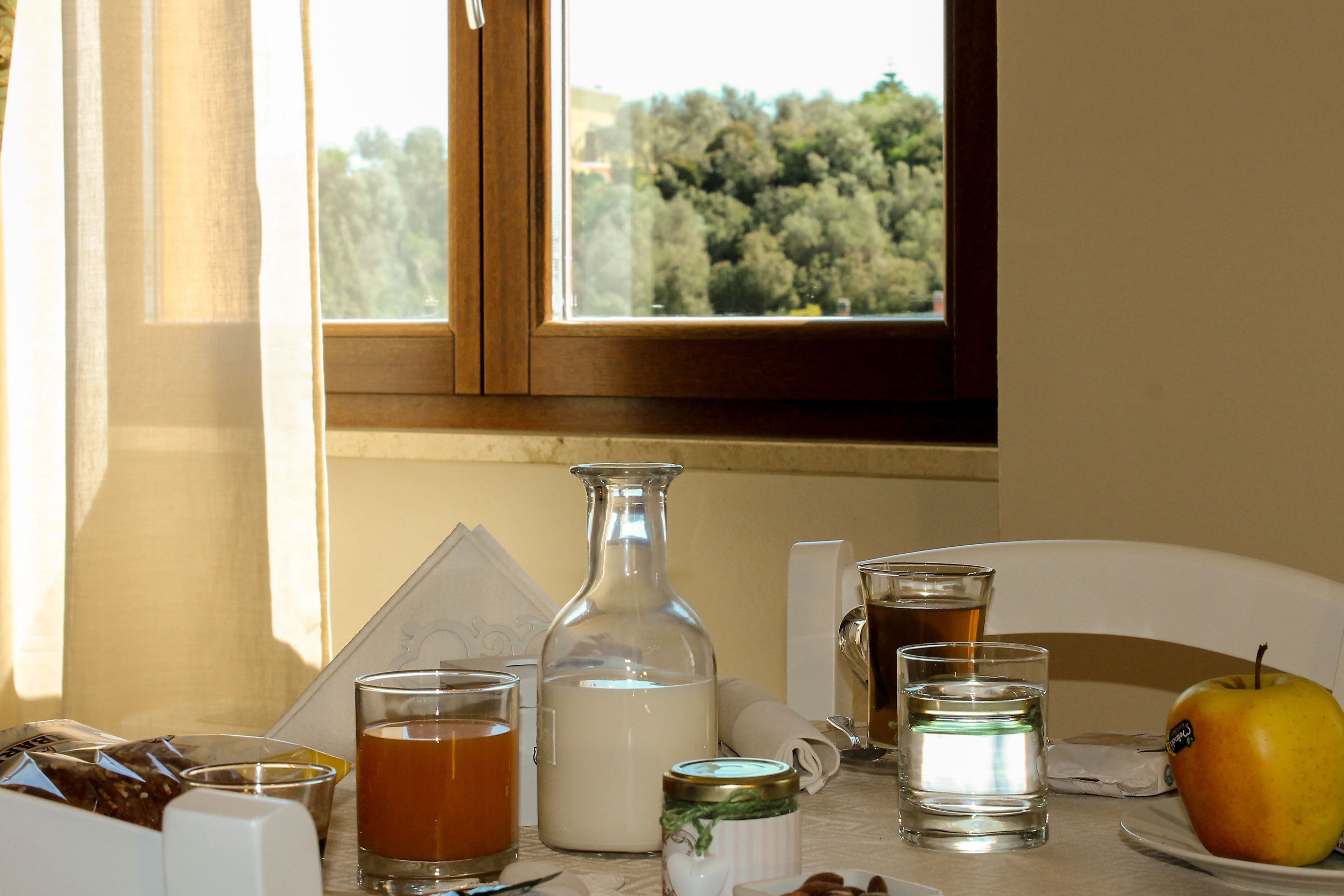 Une table avec une bouteille, des verres, un bocal, une assiette avec une pomme et une fenêtre donnant sur des arbres.