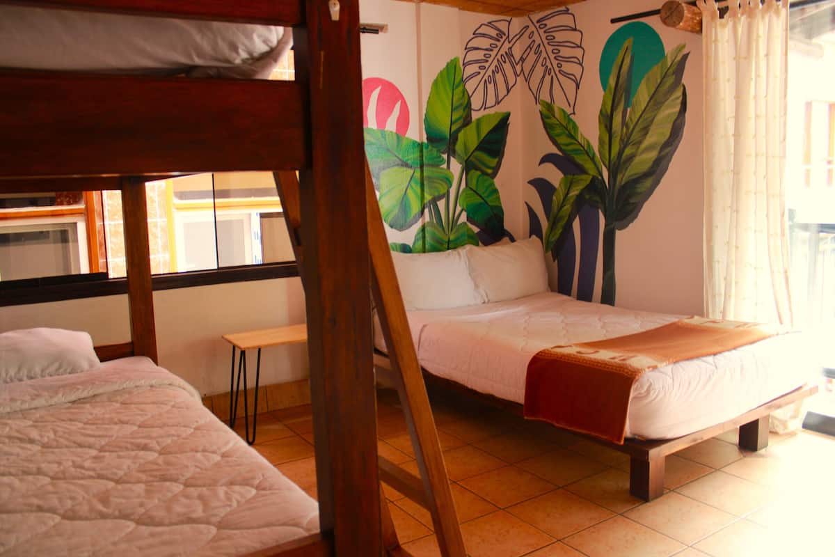 A room with two beds, a wooden headboard, a mural of tropical leaves, and a window with curtains.