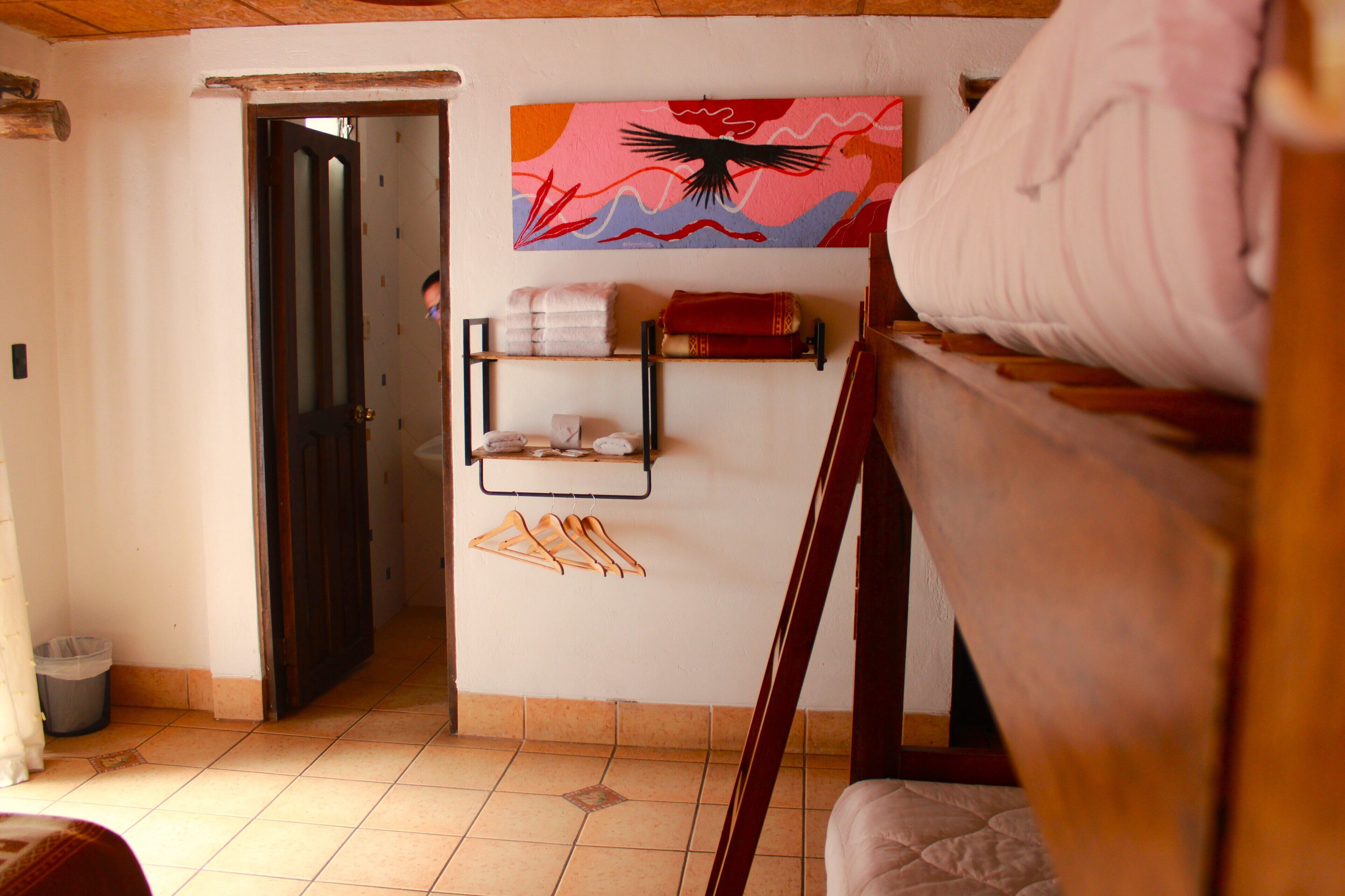 A room with a bed, a door, a wall-mounted shelf with towels, and a colorful painting.
