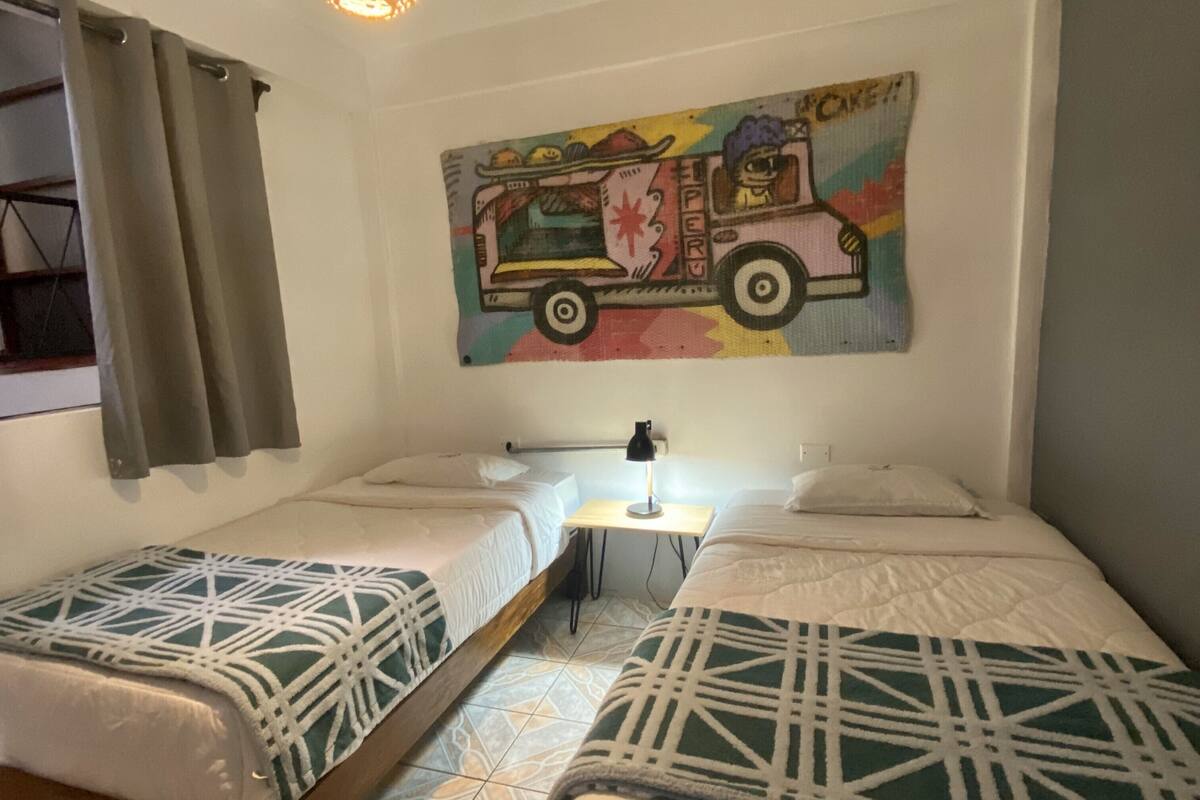 A room with two beds, a table, a lamp, and a colorful wall art of a food truck.