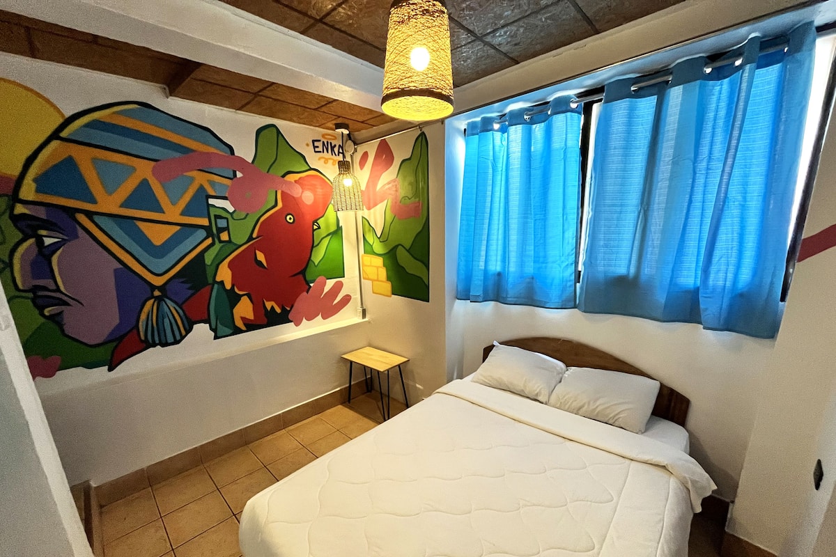 A bedroom with a bed, a small table, a colorful mural, and a window with blue curtains.