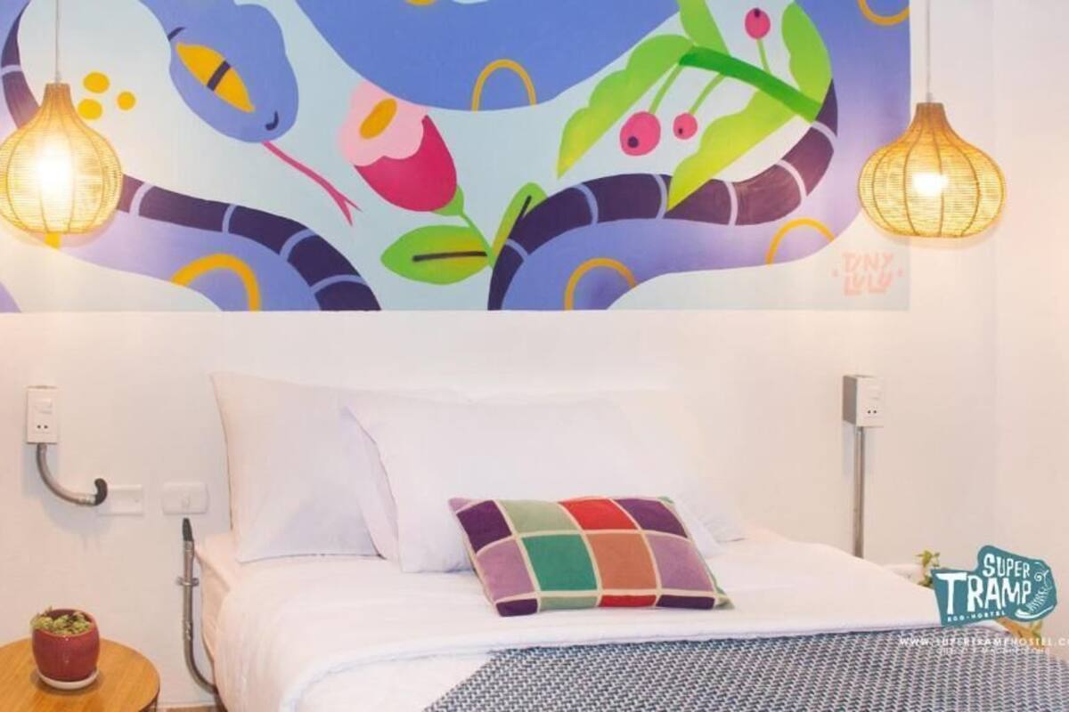 A hotel room with a bed, a colorful wall mural, two hanging lamps, and a small table with a plant.
