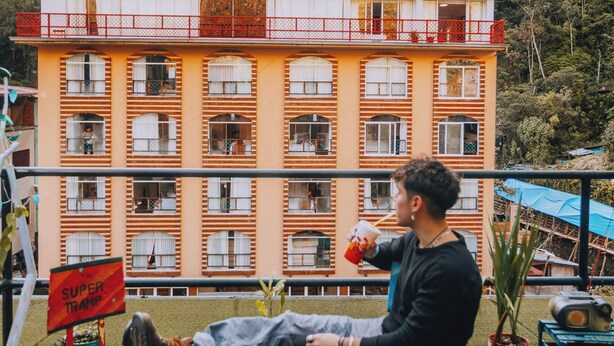 A man sitting on a colorful stool on a rooftop terrace with a view of a multi-story building and mountains in the background. - Supertramp Hostel Machupicchu (Machu Picchu)