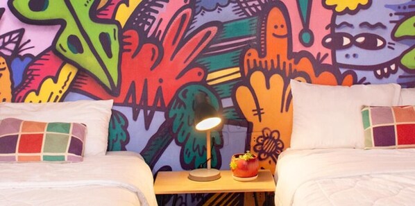 Habitación de hotel con dos camas, un cuadro abstracto de colores en la pared, una mesita de noche con lámpara y un pequeño adorno decorativo.