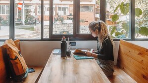 A person seated at a wooden table with a laptop, a thermos, and a bag on a bench. Large windows offer a view of buildings and trees. - Supertramp Hostel Machupicchu (Machu Picchu)