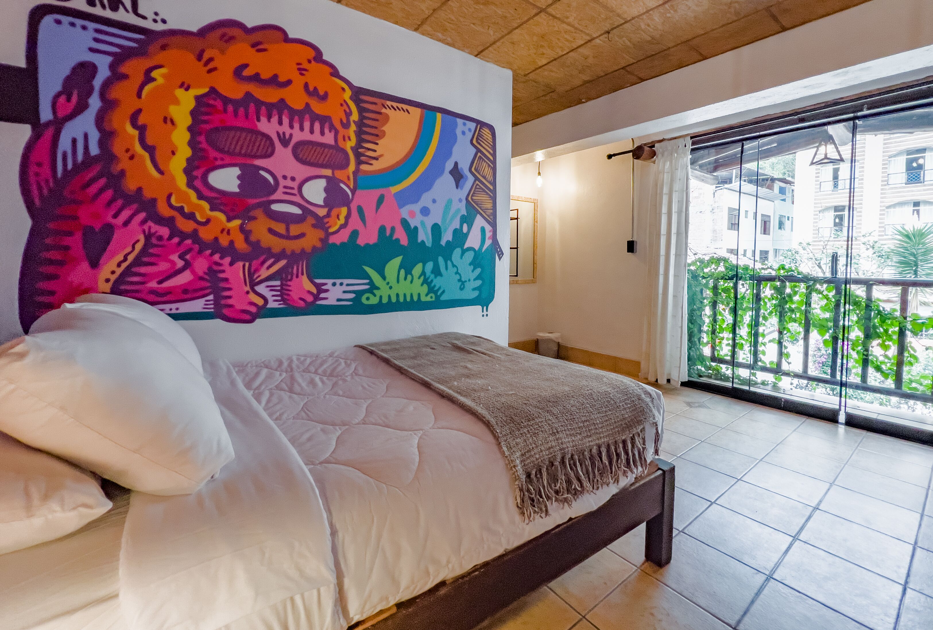 A bedroom with a large bed, a colorful mural on the wall, and a balcony with a view of buildings and greenery.