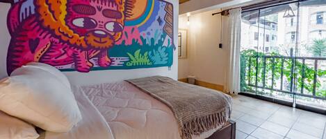 A bedroom with a large bed, a colorful mural on the wall, and a balcony with a view of buildings and greenery.