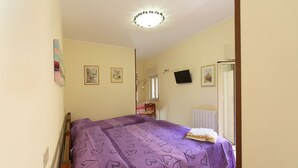 Een slaapkamer met een bed bekleed met een paarse deken met hartjes, een nachtkastje met een lamp, en een raam met uitzicht op een gebouw.