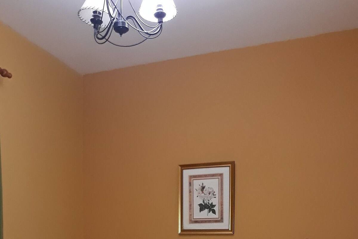 Un dormitorio con cama de madera, colcha estampada verde, dos mesitas de noche con lámparas, un cuadro en la pared y una silla de madera.