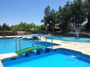 A swimming pool with a diving board, a fountain, and a surrounding area with trees and a fence. - Cabañas Bungalows Camping La Vera (Jarandilla de la Vera)