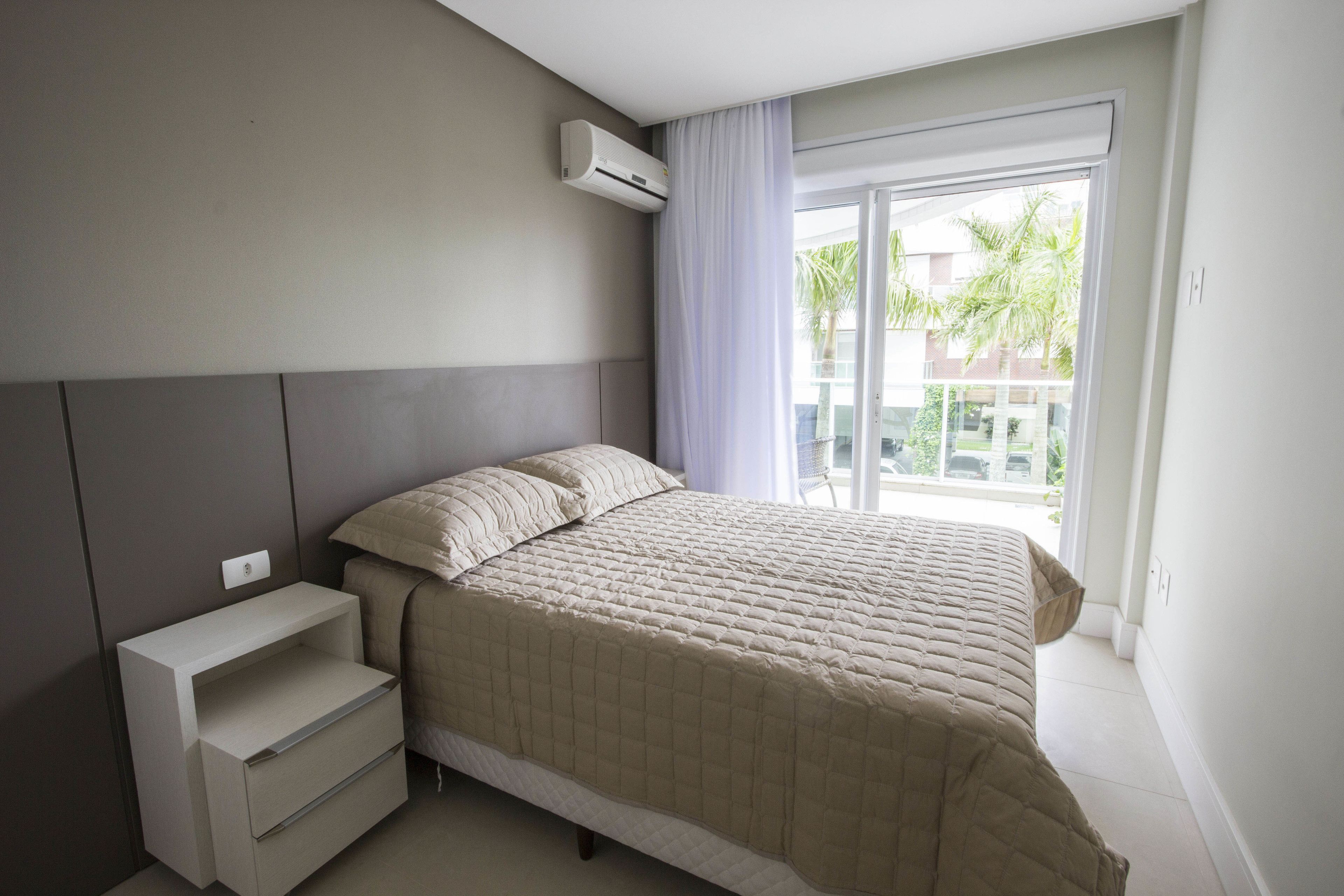 A bedroom with a bed, a nightstand, a built-in shelf, and a window with a view of palm trees and a building.