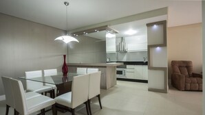 Une cuisine moderne avec des meubles blancs, un coin repas doté d’une table en verre et de chaises blanches, ainsi qu’un fauteuil marron.