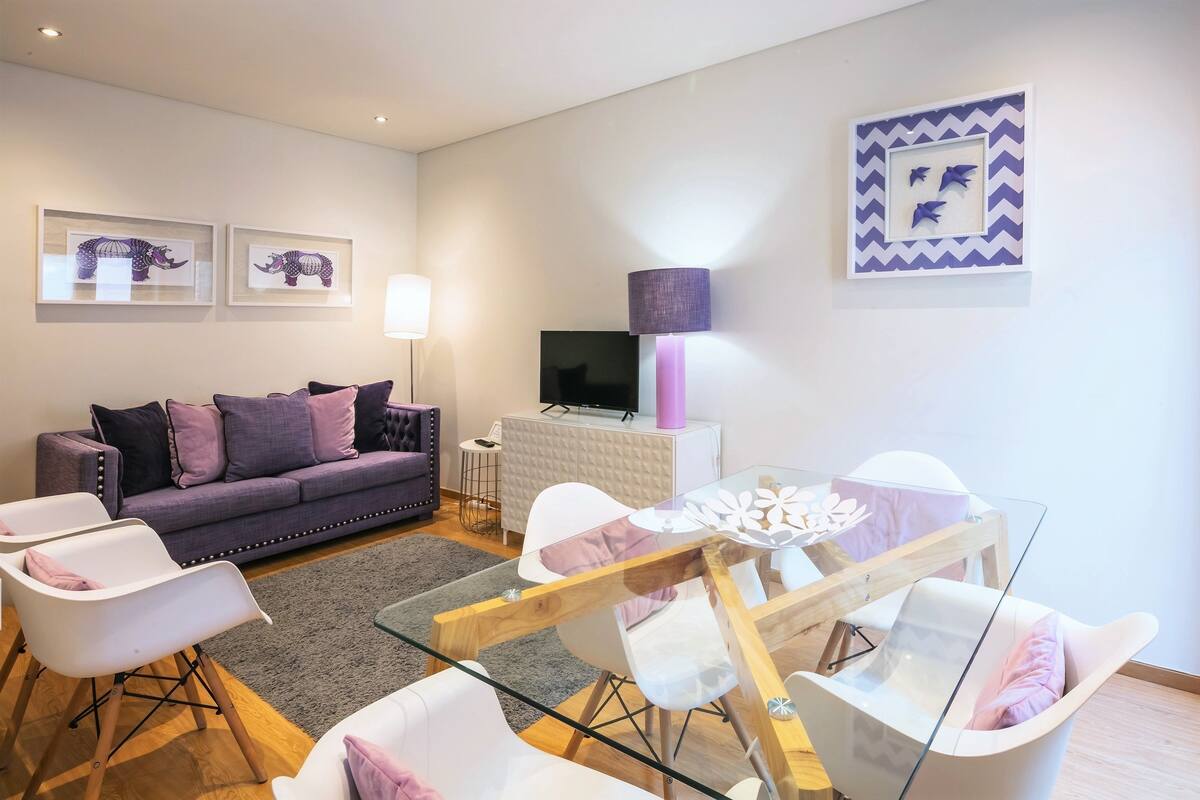 A modern living room with a sofa, chairs, a glass dining table, and artwork on the walls.