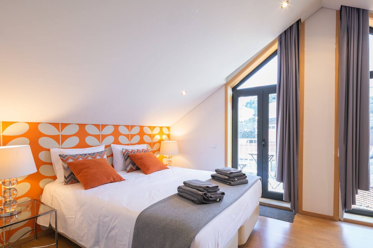 A modern hotel room with a large bed, two bedside tables with lamps, a view through a large window, and a patterned wall.