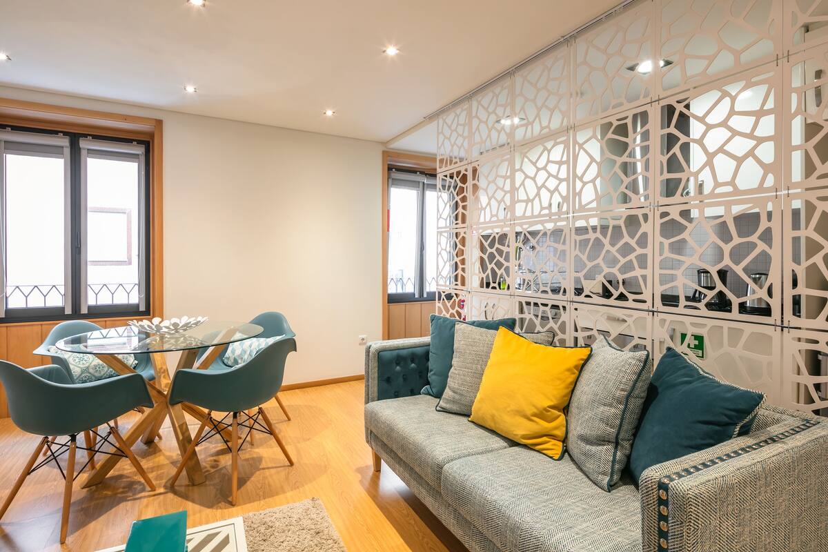 A modern living room with a glass-top dining table, teal chairs, a patterned sofa with yellow and teal cushions, and a decorative white partition.