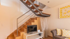 A modern living room with a wooden staircase, a flat-screen TV on a white console, a grey sofa with patterned cushions, and a decorative wall piece. - North Star Flats - Ribeira & Cellars (Vila Nova de Gaia)
