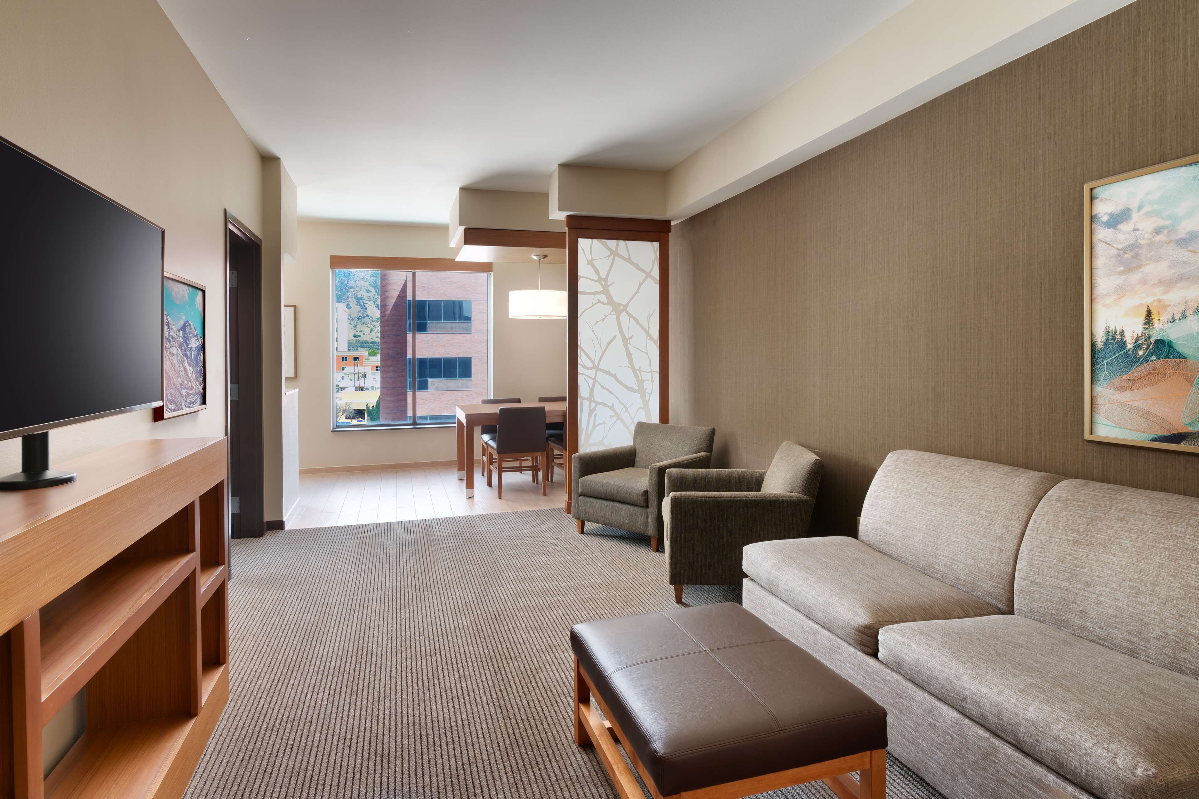 A modern hotel room with a flat-screen TV, a sofa, a coffee table, and a dining area with chairs and a table.