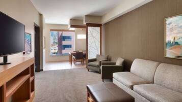 A modern hotel room with a flat-screen TV, a sofa, a coffee table, and a dining area with chairs and a table.