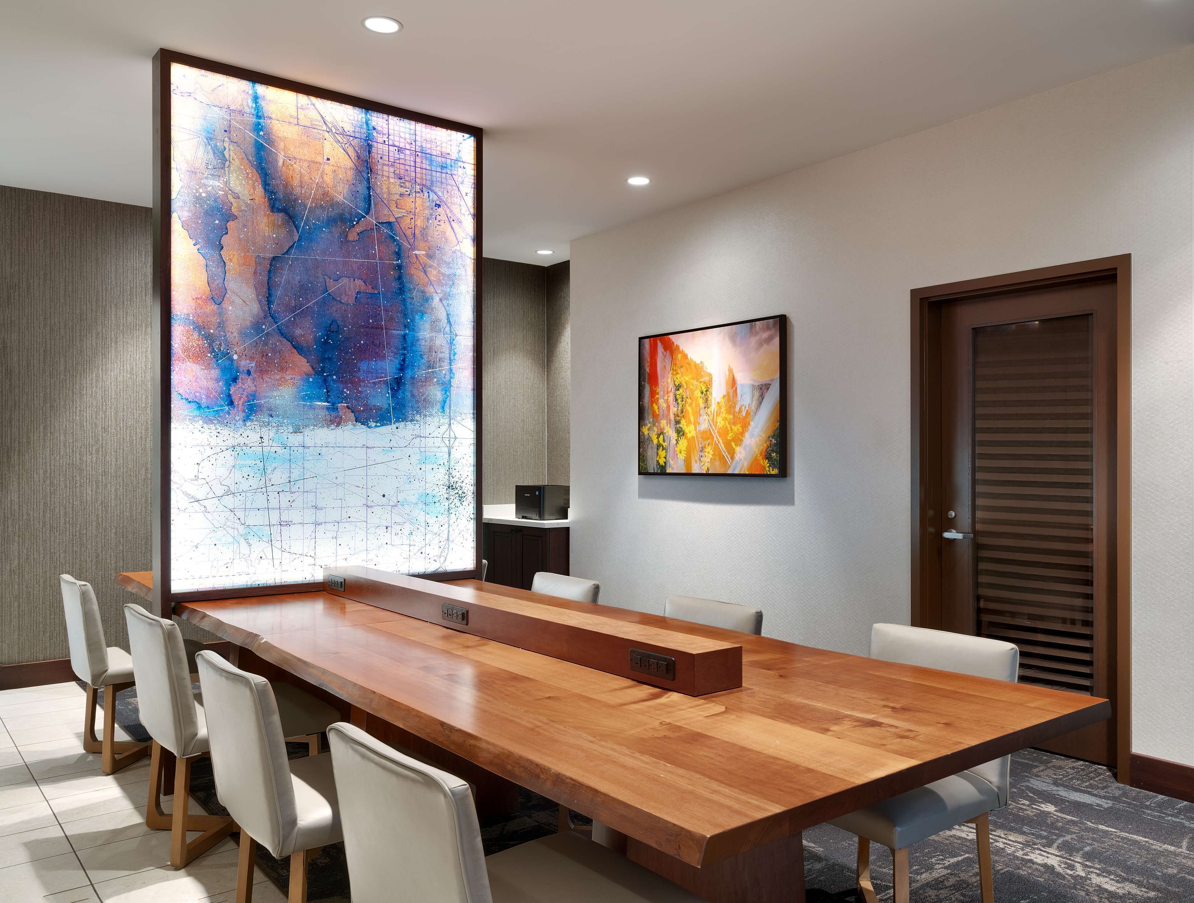 A conference room with a long wooden table, white chairs, a large abstract painting, a smaller painting, and a door with blinds.