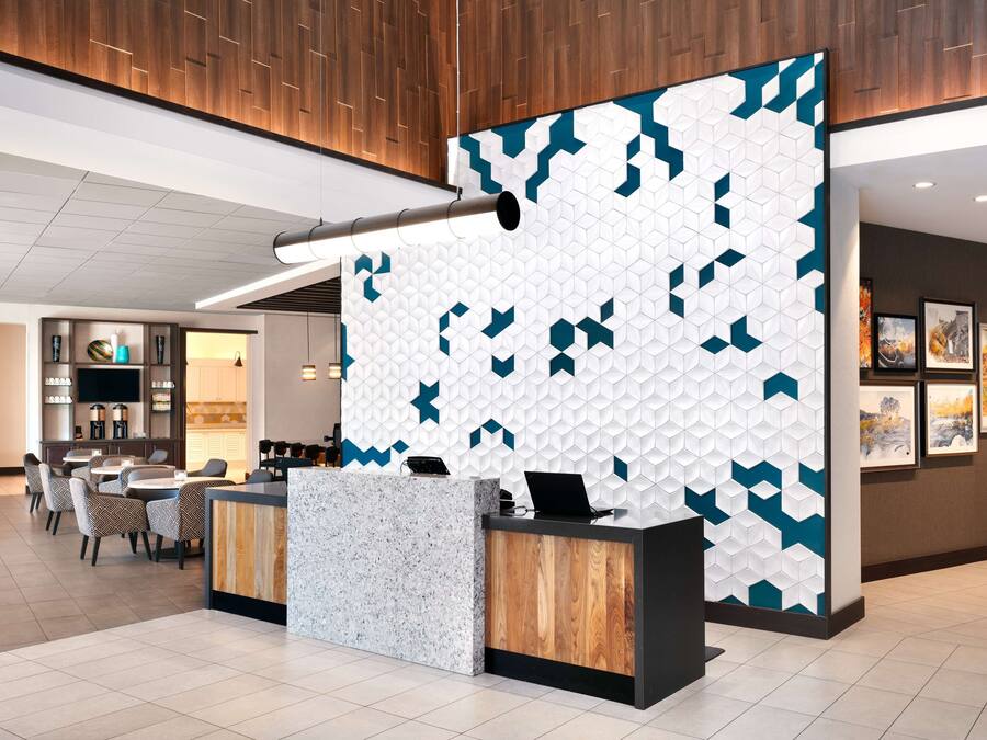 A hotel reception area with a modern design, featuring a geometric patterned wall, a reception desk with a wooden accent, and a dining area with chairs and tables.