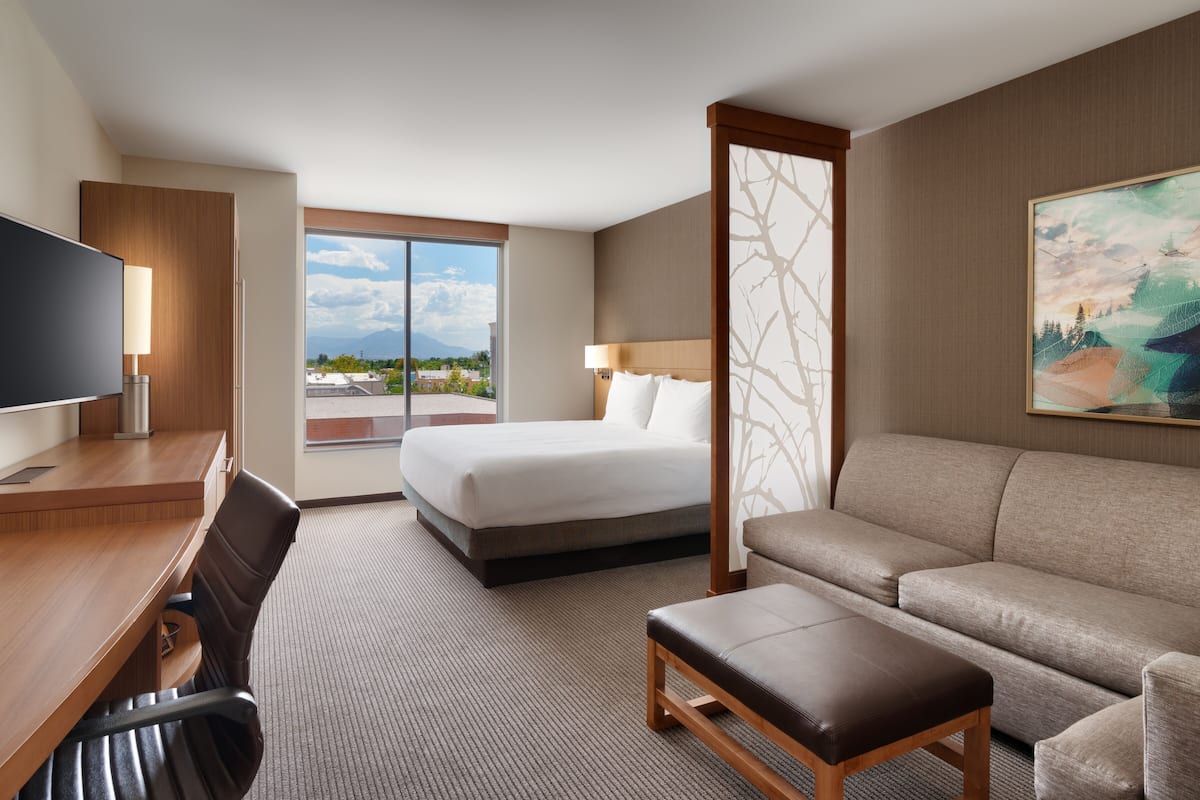 Ein Hotelzimmer mit Bett, einer Sitzecke, einem Schreibtisch mit Stuhl, einem Fernseher und einem großen Fenster mit Blick auf die Stadt.