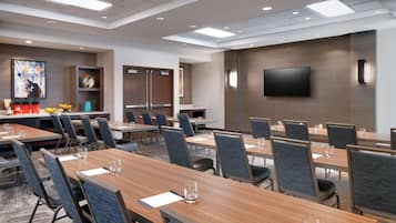 Une salle de conférence équipée de plusieurs tables et chaises, d’un téléviseur à écran plat fixé au mur, et d’un espace d’accueil agrémenté d’œuvres d’art et d’un comptoir.
