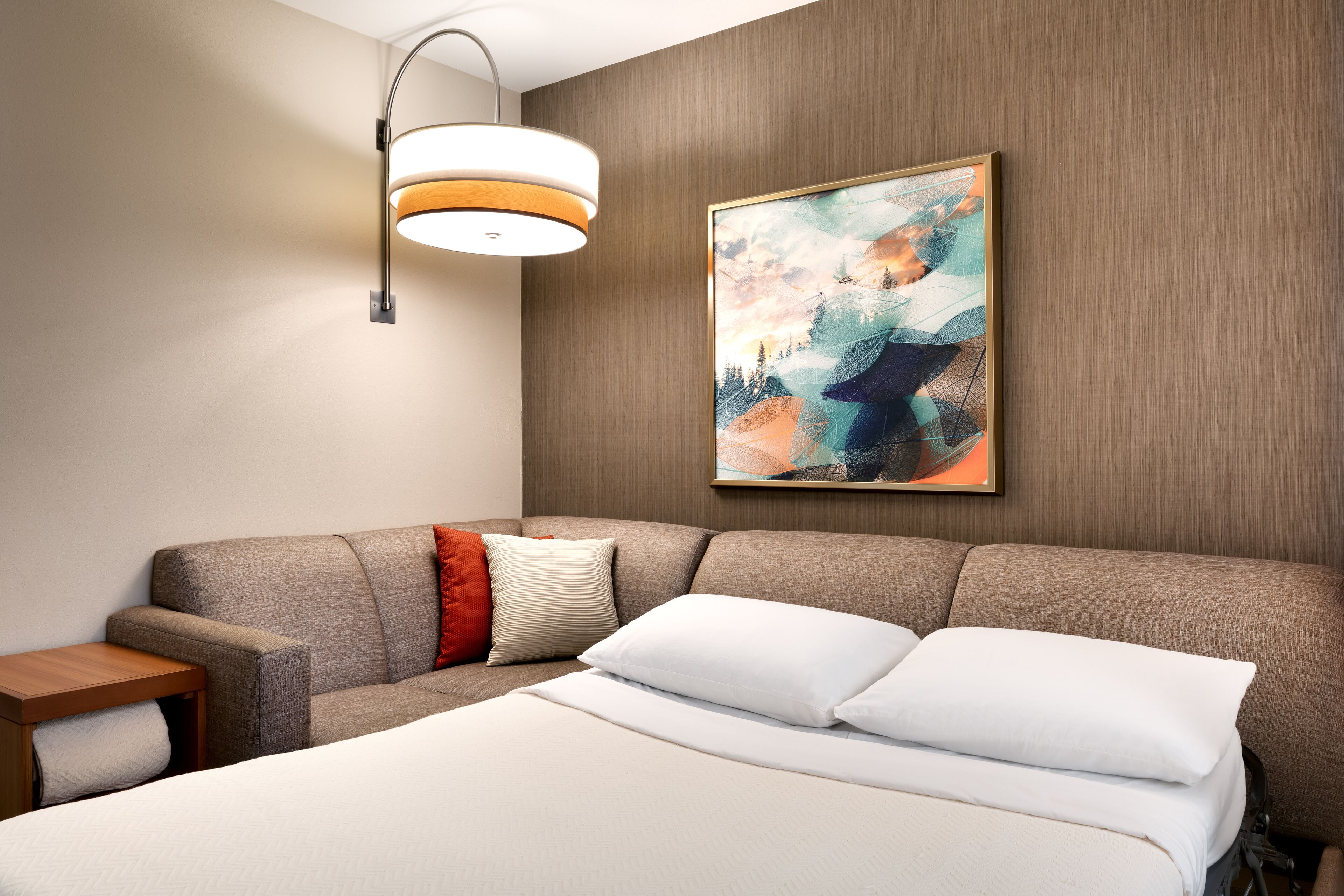 A modern hotel room with a large bed, a sofa, a small table, and a wall-mounted lamp.