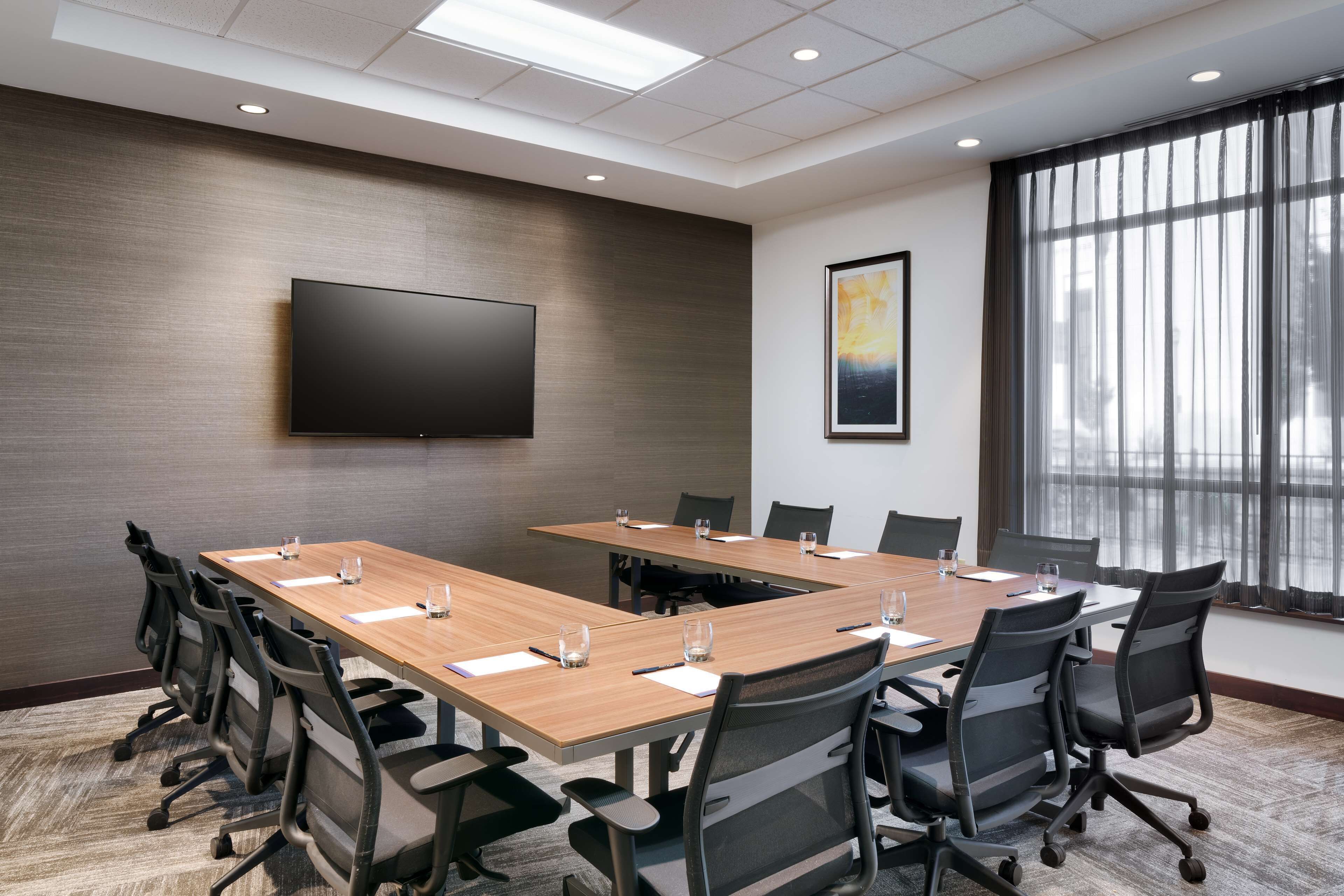 A conference room with a long table, chairs, a flat-screen TV, and a large window with curtains.