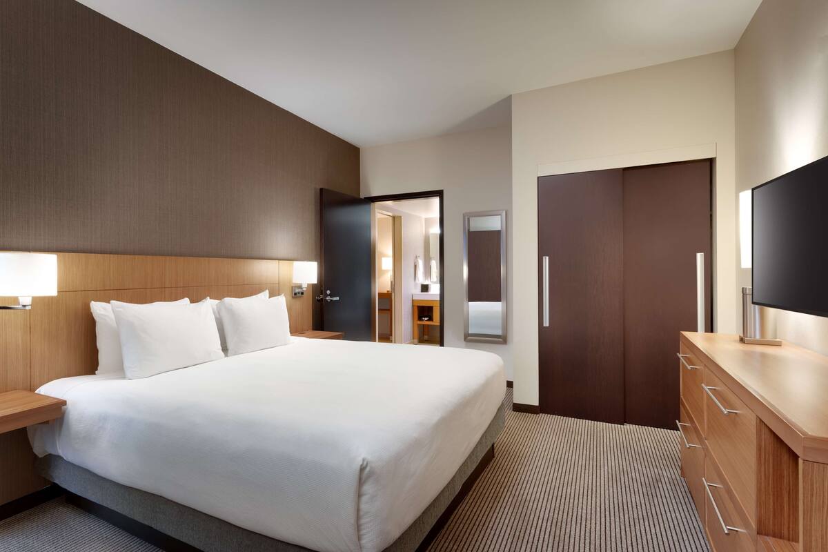 Ein Hotelzimmer mit einem großen Bett, einem Holz-Kopfteil, einem Flachbildfernseher, einem Schminktisch und einer Tür zum Badezimmer.