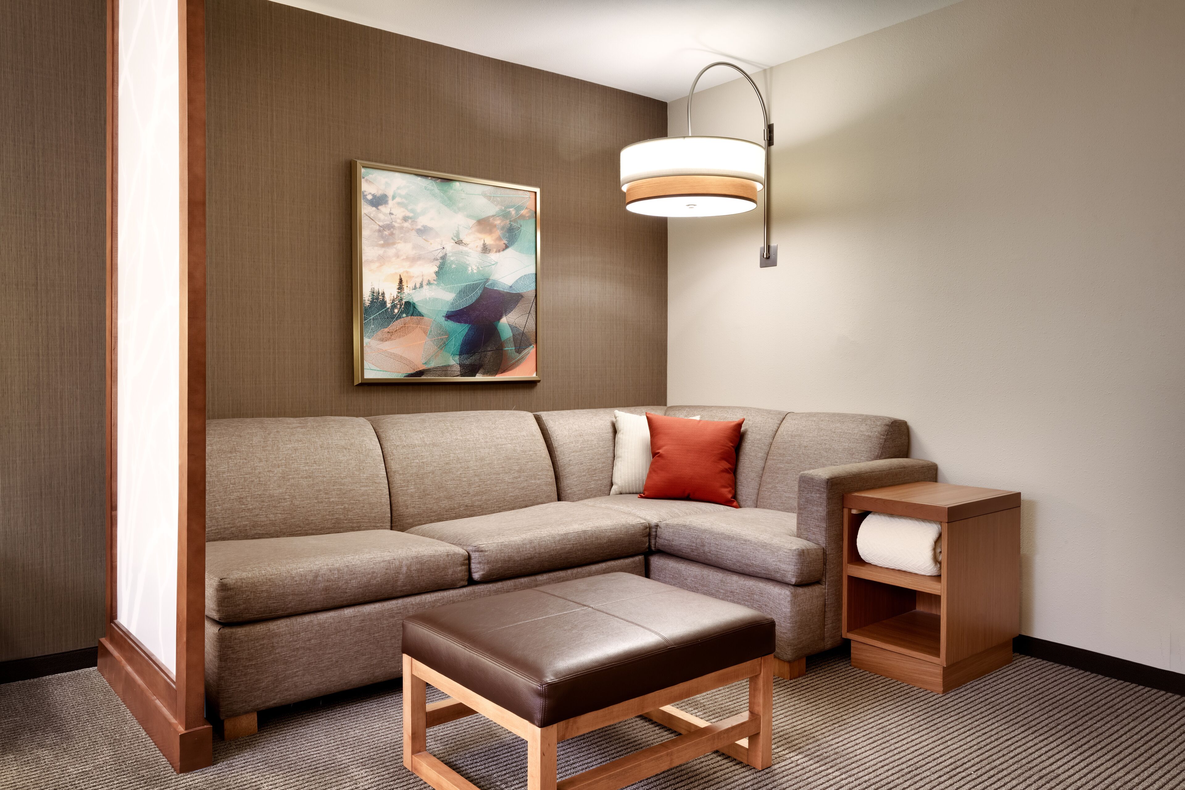 A modern living room with a sectional sofa, a wooden ottoman, a side table with a lamp, and a painting on the wall.
