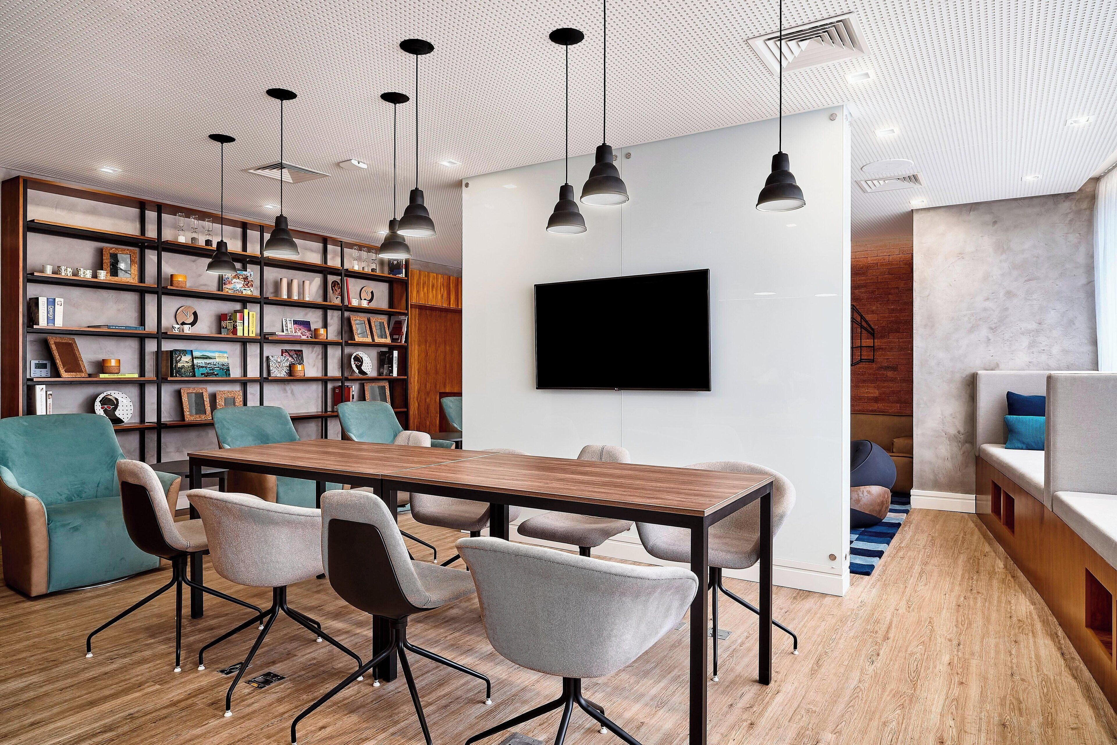 Une salle de conférence moderne, dotée d’une table en bois, de chaises rembourrées de couleur grise, d’une bibliothèque et d’un téléviseur fixé au mur.