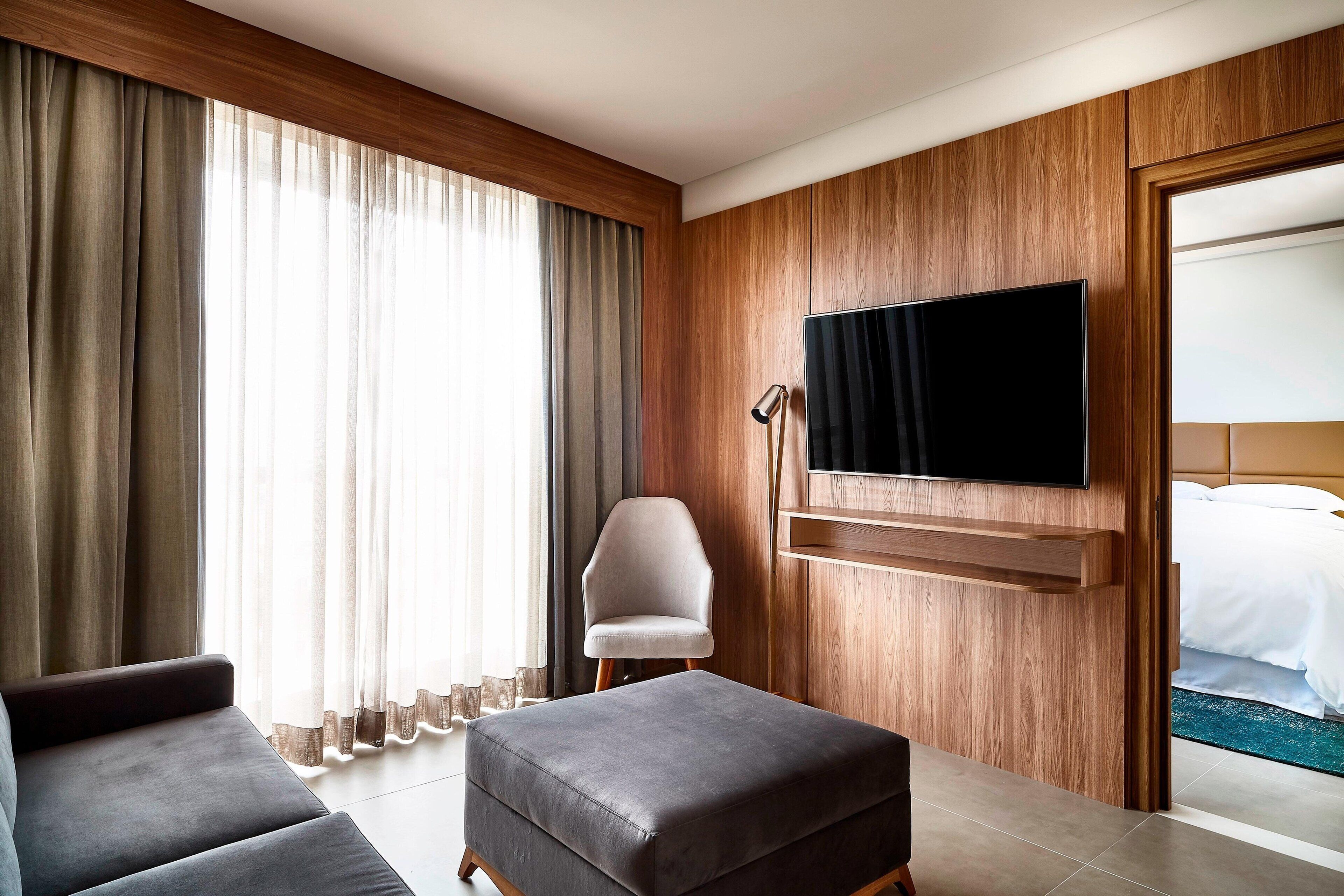 A hotel room with a large window, a flat-screen TV mounted on the wall, a grey sofa, a wooden panel wall, and a bed with white bedding.